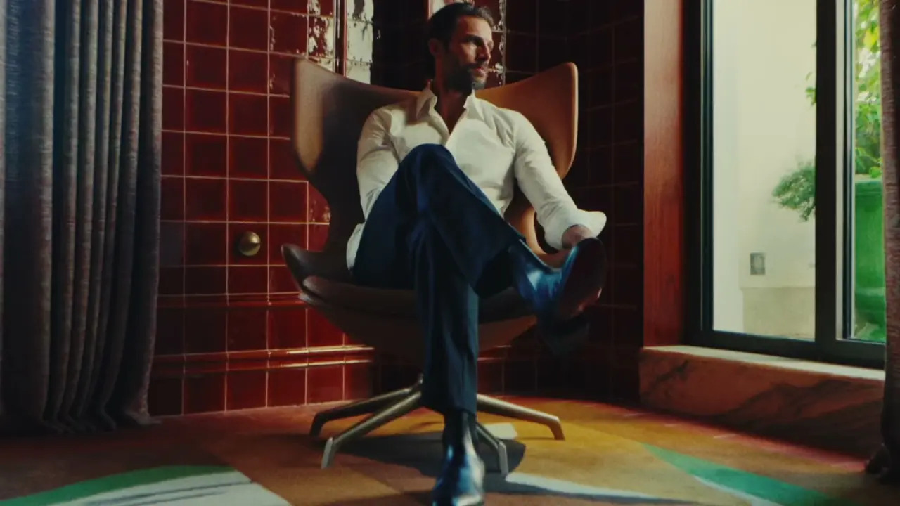 A man in a white shirt and navy trousers reclining in a leather armchair beside a deep red tiled wall, from the House of Wunder brand film